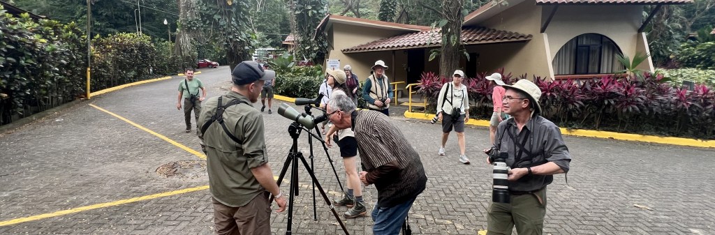 A group of photographers, some with binoculars and cameras, gather outside a building in a lush green area, preparing for a wildlife observation.