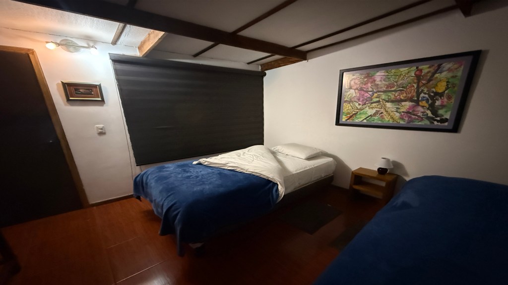 A cozy bedroom featuring two single beds with blue blankets, a nightstand, and a colorful wall art piece. The room has wooden beams and a roll-up blackout window shade.