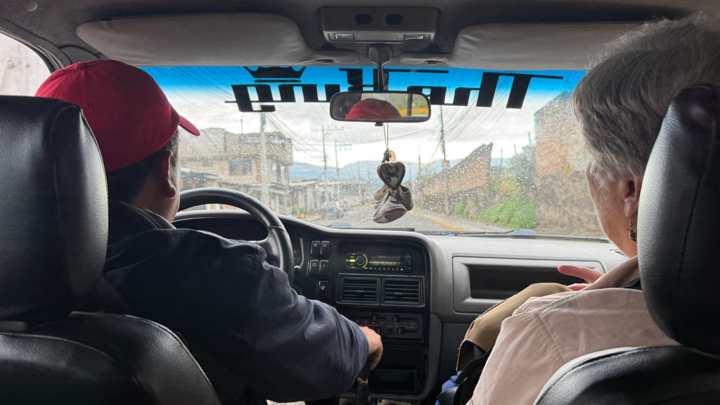 Interior view of a vehicle with a driver wearing a red cap and a passenger seated beside, looking out the window on a rainy day.