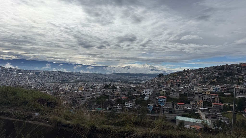 A panoramic view of a city with densely packed buildings, set against a backdrop of mountains and a cloudy sky.