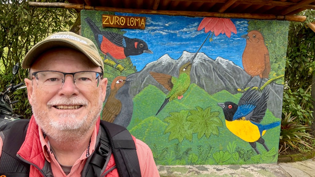 A man with glasses and a cap smiles in front of a colorful mural depicting various birds and mountains, with a sign that reads 'Zuro Loma' at the top.