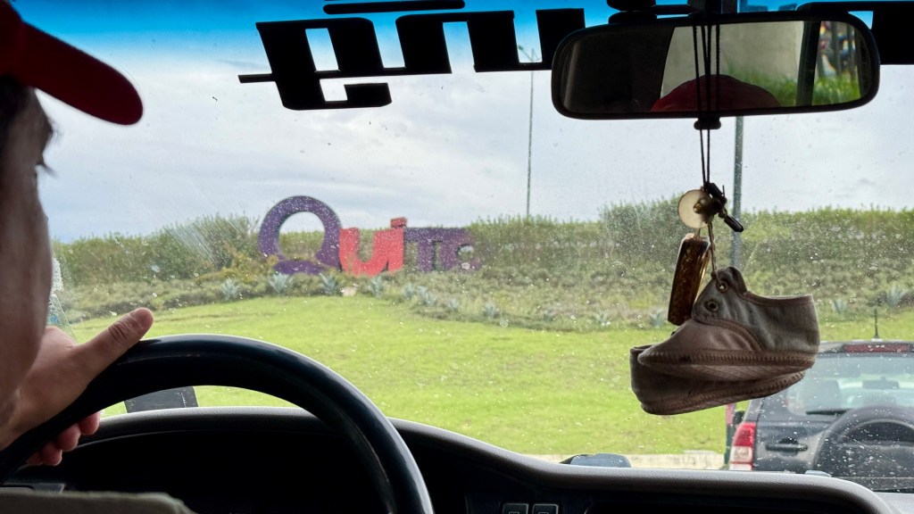 View from the driver's seat of a vehicle, showing a colorful sign that says 'Quito' in the distance, with a hanging shoe and car's steering wheel in the foreground.