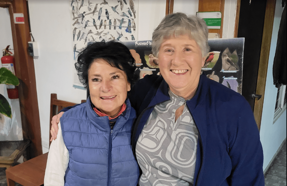 Two women pose together smiling, one in a blue vest and the other in a blue jacket, in a cozy indoor setting with bird illustrations in the background.