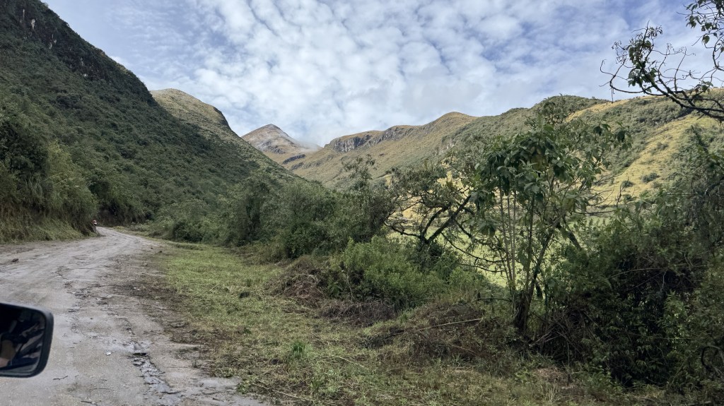 A winding dirt road surrounded by lush greenery and hills under a partly cloudy sky.