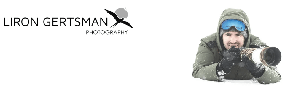 A photographer dressed in warm clothing, lying in the snow and aiming a camera with a large lens, showcasing a snowy landscape.