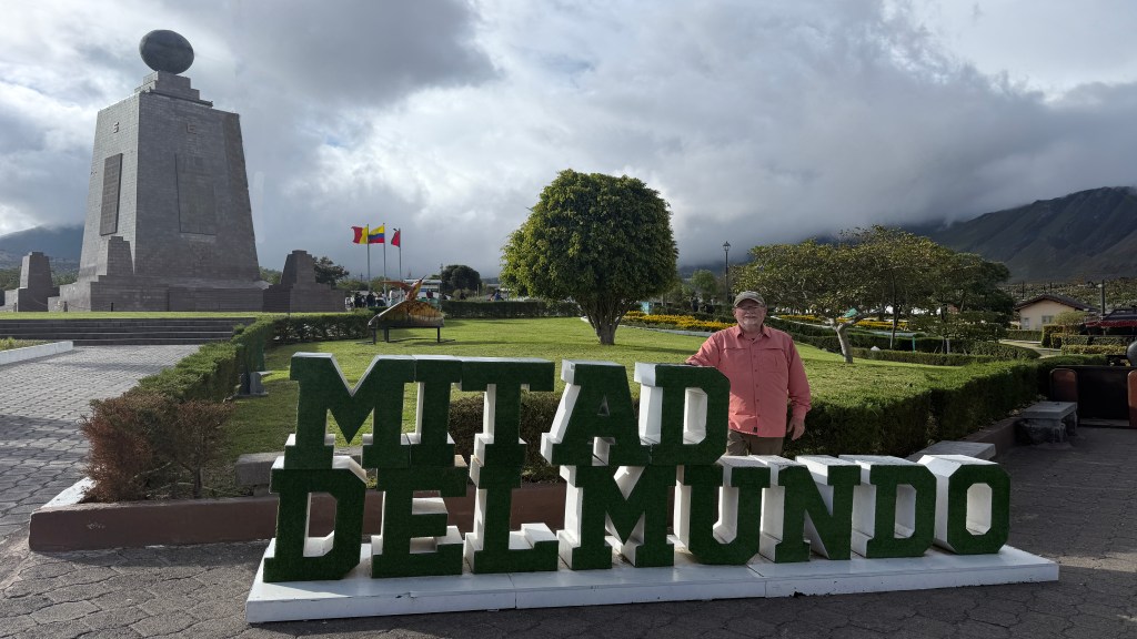 A visitor stands next to the green and white sign reading 'MITAD DEL MUNDO' in front of a monument at the Equator, with flags and mountains in the background.