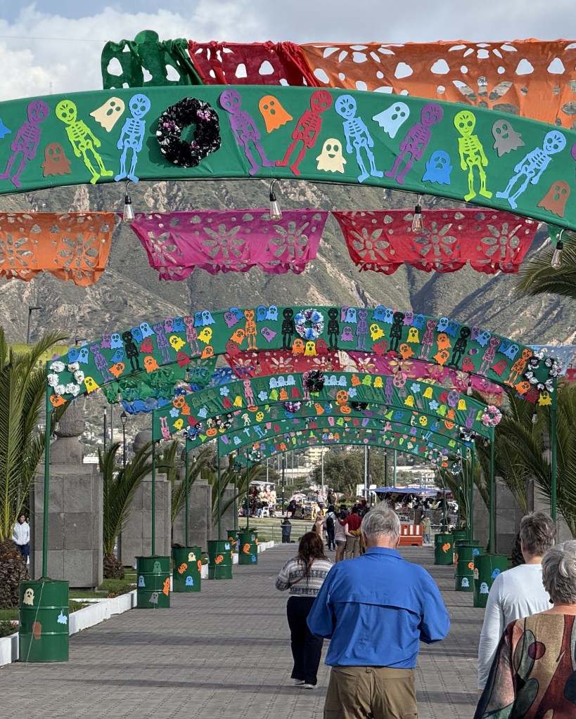 A vibrant walkway decorated with colorful papel picado banners featuring skeletons and ghosts, with people strolling underneath and palm trees lining the sides.