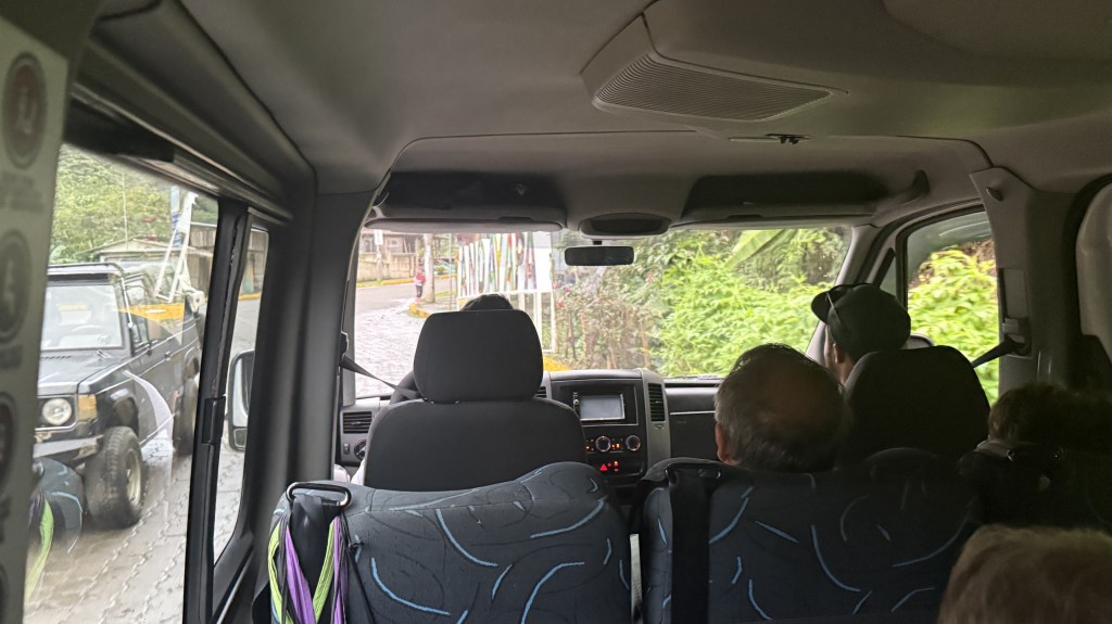 View from inside a van showing passengers and the road ahead, with green foliage visible outside and another vehicle on the side.