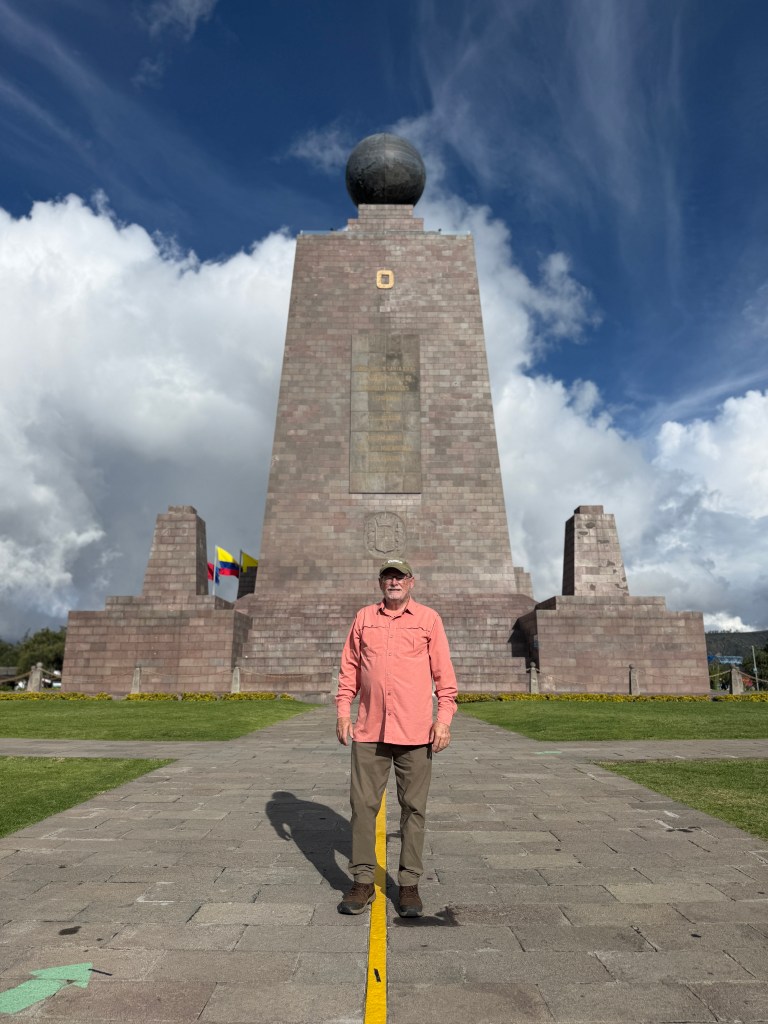 A man stands in front of the Equator Monument in Ecuador, with a globe on top and cloud-filled blue sky in the background.