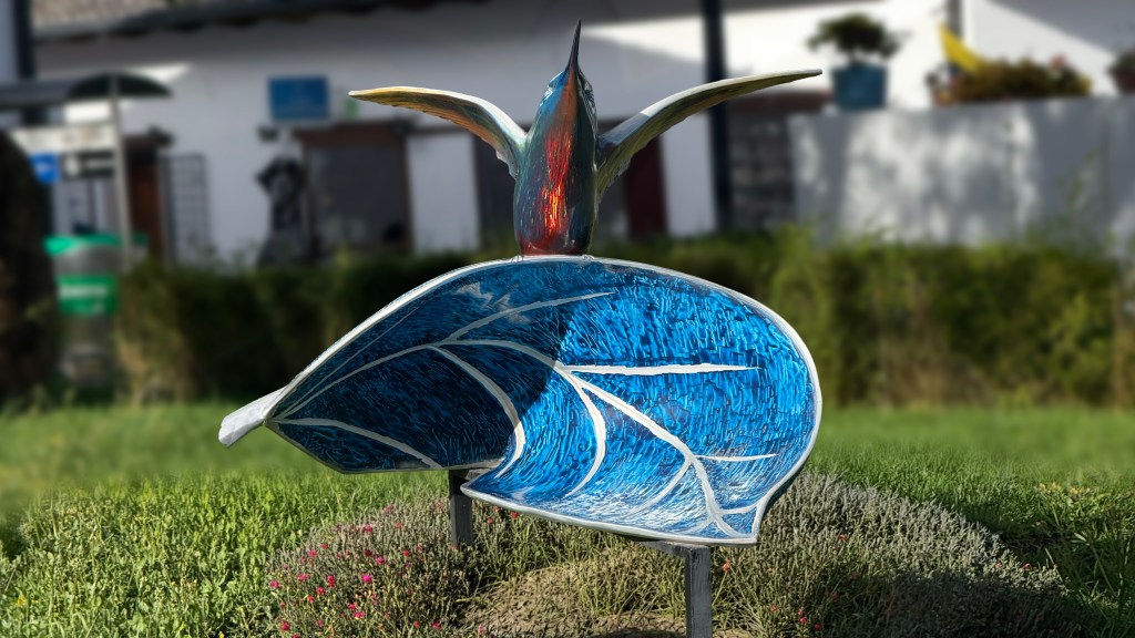 A colorful sculpture depicting a bird in flight perched on a leaf, with intricate details and vibrant blue hues, surrounded by greenery.