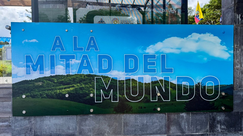 A colorful sign that reads 'A LA MITAD DEL MUNDO' against a backdrop of blue sky and green hills, indicating a landmark or tourist destination.