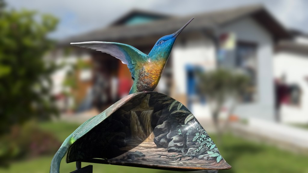 A colorful hummingbird sculpture perched on a decorative element, set against a blurred backdrop of a house and garden.