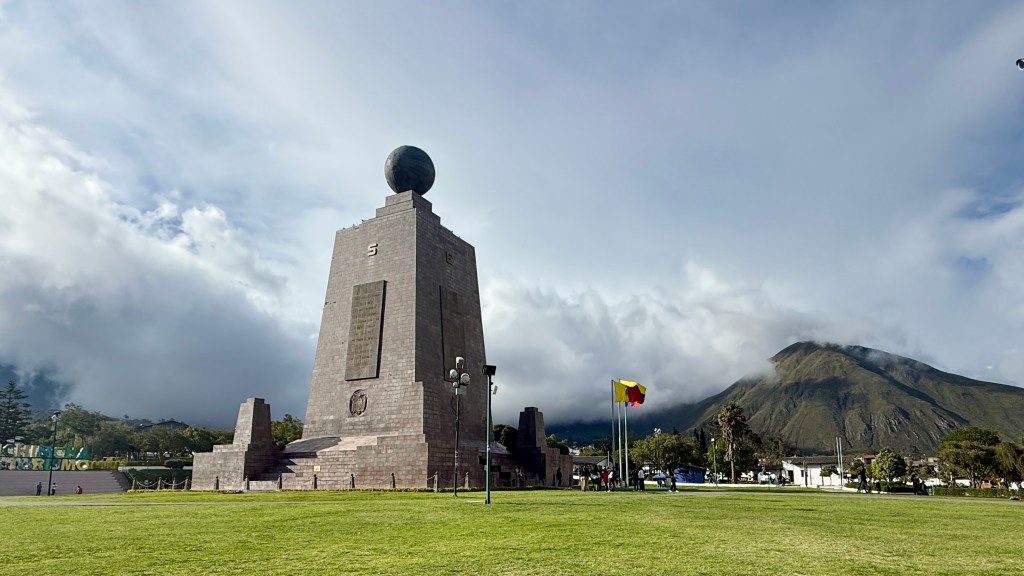 A large monument with a globe on top, located in a park surrounded by green grass and a mountain backdrop under a partly cloudy sky.